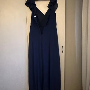 Marinblå långklänning från Bubbleroom - Säljer en marinblå långklänning från Bubbleroom med v-ringning och volangdetaljer vid axlarna. Klänningen har en elegant drapering framtill och är perfekt för festliga tillfällen. Stängs med dragkedja i bak.