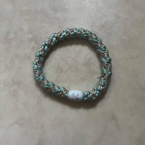 Flätat armband i blått och guld  - Säljer ett flätat armband i blått och guld med flätad design. Armbandet har ett vitt lås och är gjort av slitstark tråd. Perfekt accessoar för en personlig stil. 