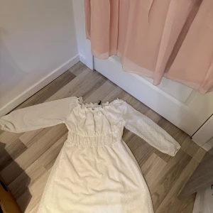 Vit långärmad klänning med resår i midjan - Söt vit klänning med långa ärmar och resår i midjan. Klänningen har en lätt struktur i tyget och en rynkad, elastisk halsringning som ger en romantisk känsla. Perfekt för sommarens tillfällen!