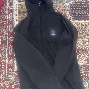 Svart jacka från Haglöfs - Säljer en svart jacka från Haglöfs med hög krage och dragkedja framtill. Jackan har en stilren design med diskret logga på bröstet och praktiska fickor med dragkedja. Perfekt för utomhusaktiviteter.