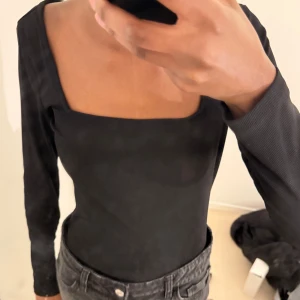 Svart långärmad bodysuit med fyrkantig ringning - Säljer en svart långärmad bodysuit med snygg fyrkantig ringning. Den har tight passform och är tillverkad i ett ribbat material som ger en stilren look. Perfekt att styla med jeans eller kjol.