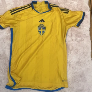 Svensk landslagströja från Adidas - Gul kortärmad fotbollströja från Adidas med blå detaljer och svenska landslagets emblem på bröstet. Tröjan har svarta Adidas-ränder på axlarna och blå kant längs sidorna och kragen. Perfekt för match eller träning. Helt äkta