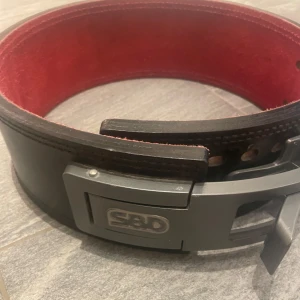 Svart träningsbälte med röd insida från SBD - Säljer ett svart träningsbälte från SBD med röd insida och kraftigt metallspänne. Bältet är brett och robust, perfekt för styrketräning och tyngdlyftning. Storlek MEDIUM