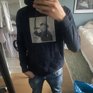 Svart hoodie med tryck från Emn - Svart hoodie från Emmamalena med ett tryck av en person som dricker ur en flaska på bröstet. Tröjan är tillverkad i mjukt material. Perfekt för en avslappnad stil.