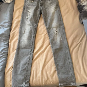 Grå slitna jeans från SMOG X-TREME - Säljer ett par grå jeans från SMOG X-TREME med slitningar och hål på flera ställen. Jeansen har en klassisk femficksdesign och raka ben. Perfekta för en avslappnad och trendig look.