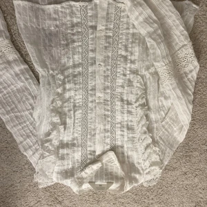 Vit blus med spets och volang - Superfin vit blus med långa ärmar, spetsdetaljer och volanger längs knäppningen. Blusen har en krage och knappar framtill, samt vackra spetsinslag på ärmarna. Perfekt för dig som gillar romantisk och bohemisk stil.