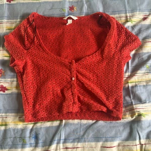 Röd croppad blus från H&M - Säljer en röd croppad blus från H&M med korta ärmar och knappar framtill. Toppen har ett hålmönster i tyget och en djup rundad urringning. Perfekt för sommardagar och enkel att styla till olika outfits.