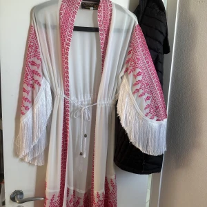 Vit och röd omlottklänning med fransar - Säljer en vit omlottklänning med röda broderade mönster och långa fransar på ärmar och nederkant. Klänningen har knytband i midjan och dekorativa detaljer längs kanterna. Perfekt för dig som vill sticka ut med en bohemisk och unik stil.