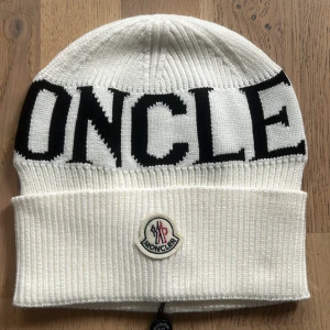 Vit stickad mössa från Moncler - Vit ribbstickad mössa från Moncler med svart logotyptext runt om och Moncler-märke framtill. Klassisk design med uppvikt kant och mjukt material som håller dig varm under kalla dagar.