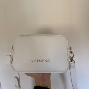 Vit axelväska från Valentino - Säljer en stilren vit axelväska från Valentino med gulddetaljer och två justerbar axelrem med logotyp och vanligt vitt skärp. Väskan har två fack med dragkedja och ett litet ytterfack med dragkedja på baksidan. Knappt använt väskan, fick inte användning för den, har kvar valentino påsarna som kom med, därav priset.