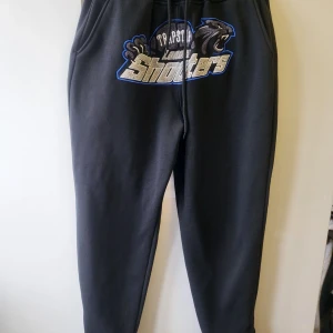  Trapstar byxor - Säljer ett par svarta mjukisbyxor från Trapstar med ett coolt tryck framtill. Byxorna har en elastisk midja med snörning och sidofickor. Perfekta för en avslappnad stil.