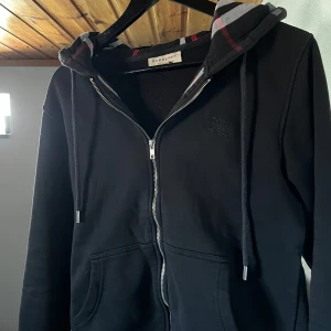 Svart hoodie från Burberry - Säljer en stilren svart hoodie från Burberry med dragkedja och broderad logga på bröstet. Den har en klassisk rutig insida av huvan i grått och rött. Perfekt för en avslappnad och trendig look.