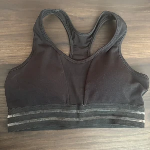 Svart sport-bh - Säljer en svart sport-bh med brottarrygg och elastiskt band med grå ränder. Perfekt för träning och ger bra stöd. Använd 1 gång