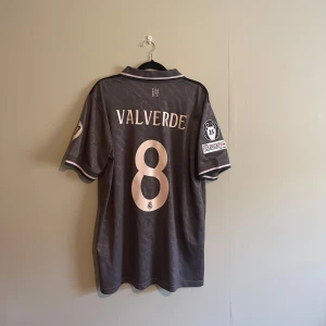 Real Madrid fotbollströja 24/25 tredjeställ - (Det står L men den passar perfekt på en M också)Säljer en grå fotbollströja från Adidas med Real Madrids emblem och sponsorlogga 'Emirates Fly Better' på framsidan. Tröjan har korta ärmar med detaljer och nummer 8 på baksidan. Perfekt för fotbollsfans!