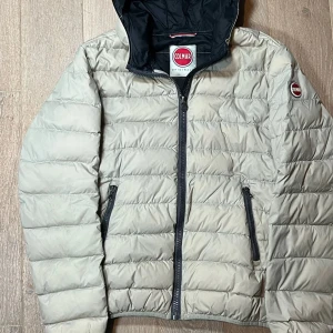 Beige dunjacka från Colmar - Säljer en snygg beige dunjacka från Colmar med huva och dragkedja. Jackan har en quiltad design och två praktiska fickor med dragkedjor. Perfekt för kyligare dagar!