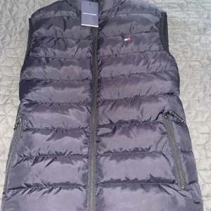 Marinblå dunväst från Tommy Hilfiger - Snygg marinblå dunväst från Tommy Hilfiger med dragkedja och diskret logga på bröstet. Perfekt för lager-på-lager under kyliga dagar. Västen har praktiska fickor med dragkedja för säker förvaring.