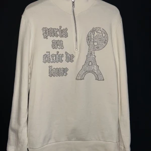 Half zip Tröja  - Säljer en vit tröja från Blinglucid med dragkedja och ett glittrigt tryck av Eiffeltornet och texten 'precis au clair de lune'. Tröjan har långa ärmar och en hög krage. Perfekt för en stilren look. Inprencip helt ny, köpte den för något år sen till en skolavslutning med den blev alldeles för stor så vi la undan den, och den passar riktigt inte min stil. Storlek Xs 