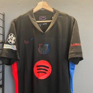 Svart fotbollströja från Nike - Säljer en svart fotbollströja från Nike med röda och blå detaljer. Tröjan har korta ärmar och ett tryck av en logotyp på framsidan. På baksidan finns namnet 'Raphinha' och nummer 11 i rött. Perfekt för fotbollsfans! Helt ny med tagg i storlek M en replika