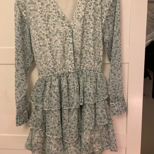 Blommig klänning med volanger - Söt blommig klänning från Gina Tricot med volanger och v-ringad hals. Klänningen har långa ärmar och ett fint blommönster i grönt och vitt. Använd ca 3 gånger💕Passar S också!