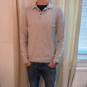 Grå pikétröja - Säljer en stilren grå pikétröja med långa ärmar och knappar vid kragen. Perfekt för en avslappnad men ändå snygg look. Tröjan har ribbade muddar vid ärmslut och nederkant.