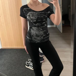 Svart topp med tryck - Snygg svart topp med ett unikt tryck av rosor och dödskallar i silver. Toppen har en lite tajtare passform och korta ärmar, perfekt för en cool och avslappnad stil. Märket är Madonna for girls! Storleken är L men jag har sytt om den så den sitter som en xs-s i midjan