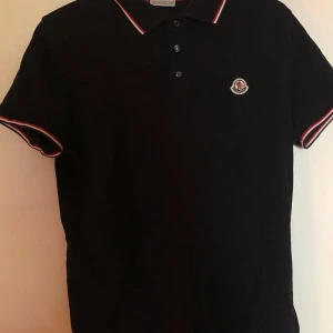 Svart pikétröja från Moncler - Snygg svart pikétröja från Moncler med klassisk krage och knappar. Tröjan har röda och vita detaljer på kragen och ärmsluten samt Monclers logotyp broderad på bröstet. Perfekt för en stilren look!