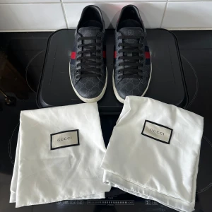Svarta sneakers med röd och blå detalj från Gucci - Snygga svarta sneakers från Gucci (Äkta) med ikoniskt mönster och röd samt blå rand på sidan. Skorna har en rund tå och snörning framtill. Baksidan har en kontrasterande röd och blå detalj med Gucci-logga. Skorna är i storlek 7 så motsvarar 42-42  och är bara använda fåtalet gånger vid speciella tillfällen. Skopåsarna följer med men tyvärr inte boxen då jag använt den till annat.
