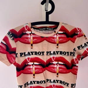 Croptop från Playboy x Missguided - Säljer en beige t-shirt från Playboy x Missguided med ett iögonfallande tryck av röda läppar och cigaretter. T-shirten har korta ärmar och en rund halsringning. Perfekt för dig som vill sticka ut!