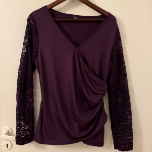 Lila omlottopp med spetsärmar  - Snygg lila omlottopp med spetsdetaljer på ärmarna. Toppen har en v-ringad design och långa ärmar, perfekt för en stilren look. Passar utmärkt till både jeans och kjol.