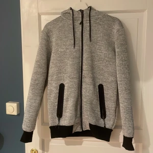 Grå och svart jacka/hoodie från Smog - Zip tröja med mjukt pälsaltigt material på insidan från Smog. Jackan har en dragkedja framtill och en mysig huva med snörning. Riktigt mysig.
