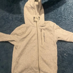 Grå huvtröja från Sail Racing - Säljer en grå huvtröja från Sail Racing med dragkedja och en bröstficka med dragkedja. Tröjan har långa ärmar och en bekväm huva med snörning. Perfekt för kyliga dagar!