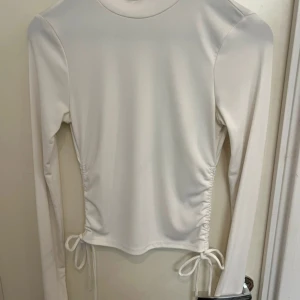 Vit långärmad topp från chiquelle - Säljer en stilren vit långärmad topp från chiquelle. Toppen har en hög krage och snörning i sidorna för en justerbar passform. Perfekt för en minimalistisk look. 