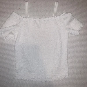 Vit offshoulder topp - Säljer en söt vit offshoulder topp med smockdetaljer och tunna axelband. Perfekt för varma sommardagar. Toppen har en luftig och bekväm passform med korta ärmar.
