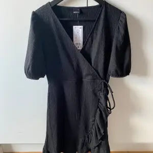 Snygg svart omlottklänning från Gina Tricot med korta ärmar och volangdetaljer. Perfekt för en stilren look. Klänningen har en v-ringad design och knytning i midjan för en smickrande passform.🩷