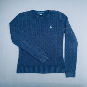 Blå stickad tröja från Ralph Lauren - Säljer en snygg blå stickad tröja från Ralph Lauren med kabelstickat mönster och broderad logga på bröstet. Tröjan har långa ärmar och ribbstickade muddar vid ärmslut. Står att den är M men passar mer som en mellanliten S