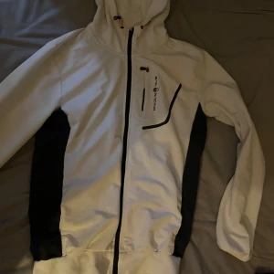 Vit jacka från Sail Racing - Snygg vit jacka från Sail Racing med svarta detaljer. Jackan har en dragkedja framtill och en huva. Perfekt för en sportig look.