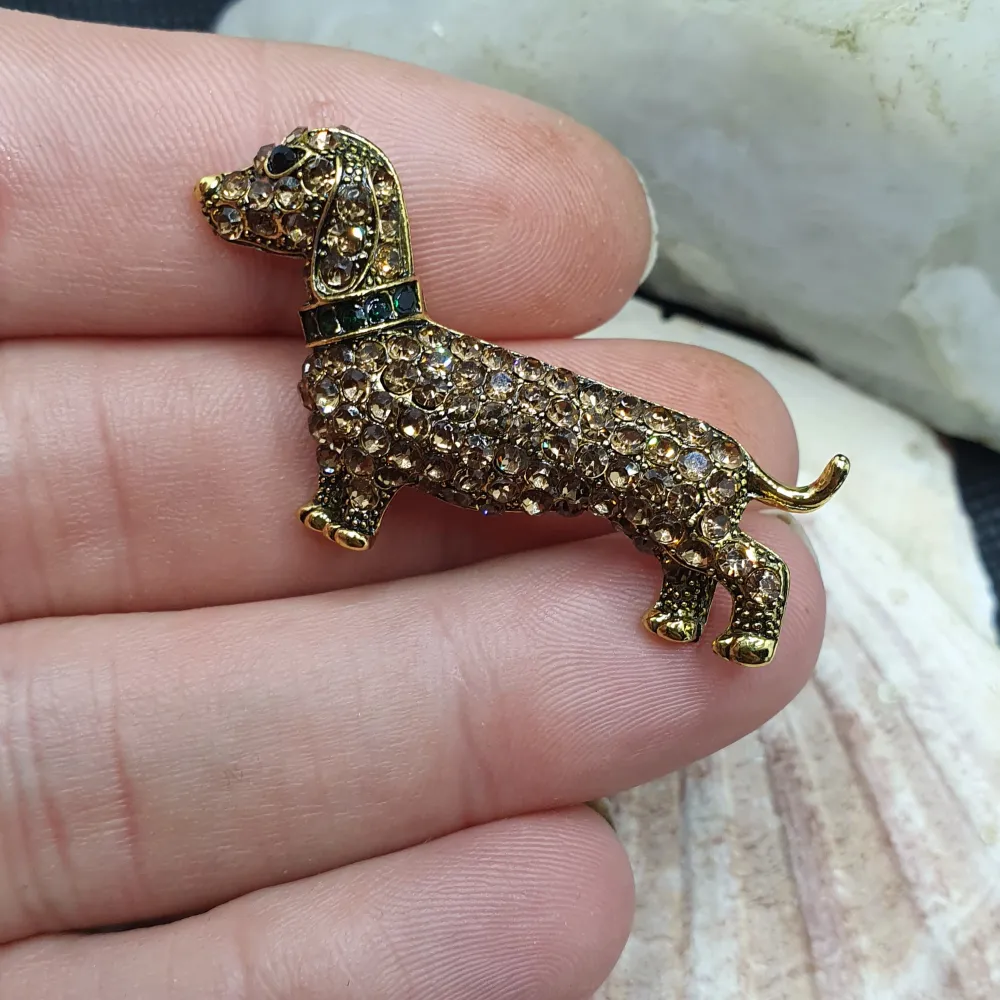 Den här broschen föreställer en tax och måtten är ca 4.5 ×2.5 cm. Guldfärgad metall med vacker ytstruktur och emaljering samt ytan täckt av kristaller. Otroligt vacker brosch. Asusteet.