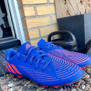 Blå och röda fotbollsskor från Adidas - Snygga blå fotbollsskor från Adidas med röda detaljer och snörning. Perfekta för fotbollsplanen med sin sportiga design och greppvänliga dobbar.