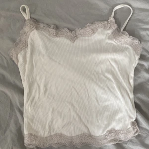 Vit ribbad linnetopp med spetsdetaljer - Säljer en söt vit ribbad linnetopp med smala axelband och spetsdetaljer längs kanterna. Perfekt för varma sommardagar eller som ett lager under en tröja.