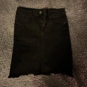 Svart jeanskjol - Snygg svart jeanskjol med klassisk femficksdesign och rå kant. Kjolen har en dragkedja och knapp framtill för enkel påklädning. Perfekt för en trendig och avslappnad stil.