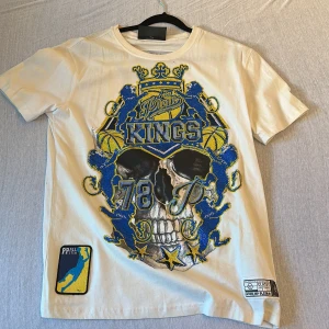 Vit t-shirt från Philipp Plein - Säljer en cool vit t-shirt från Philipp Plein med ett stort tryck av en dödskalle och texten 'Kings' i blått och gult på framsidan. Baksidan har ett PP All Star-märke i blått och gult. Perfekt för dig som vill sticka ut!
