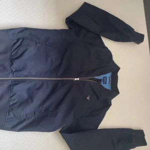 Mörkblå vår jacka från GANT - Säljer en stilren mörkblå vårjacka från GANT med dragkedja och ribbade detaljer. Jackan har en ljusblå insida med GANT-logga och är perfekt för en casual look. Vid Frågor är det bara att höra av dig!