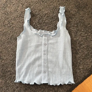 Ljusblå ribbad linnetopp - Säljer en söt ljusblå ribbad linnetopp med knappar framtill. Toppen har volangdetaljer vid axelbanden och halsringningen, vilket ger en romantisk touch. Perfekt för varma dagar!