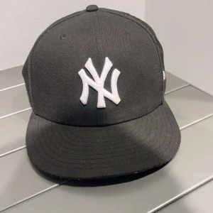 Svart keps från New Era - Snygg svart keps från New Era med vit broderad logga framtill. Perfekt för en stilren look. Kepsen har en klassisk design med böjd skärm och är en del av 59FIFTY-serien.