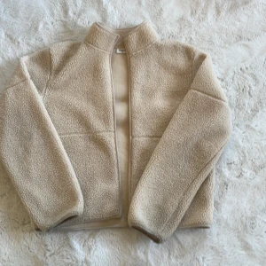 Beige teddyjacka - Mysig beige teddyjacka med hög krage och långärmad design. Perfekt för kyliga dagar med sin mjuka och varma känsla. Jackan har en enkel och stilren look utan synliga detaljer som dragkedja eller knappar. Storlek 170 barn, passar XS/S