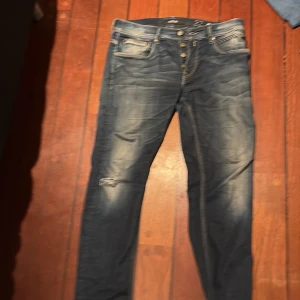 Blå jeans från Replay - Snygga blå jeans från Replay med knappar i gylfen och klassisk femficksdesign. Jeansen har en lätt tvättad look och kontrastsömmar som ger en cool stil. Perfekta för en avslappnad vardagslook.
