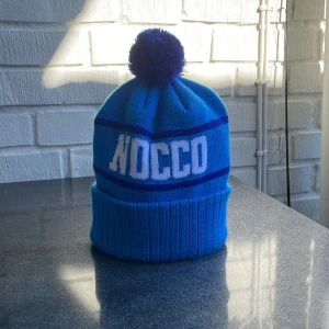 Blå mössa med tofs från NOCCO - Snygg blå mössa från NOCCO med en stor tofs på toppen. Mössan har ett ribbat uppvik och texten 'NOCCO' i vitt. Perfekt för kyliga dagar.