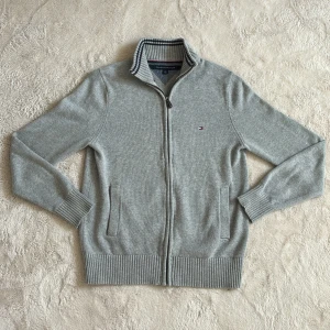 Grå kofta från Tommy Hilfiger - Snygg grå kofta från Tommy Hilfiger med dragkedja och ribbade detaljer. Den har en diskret logga på bröstet och randig krage i blått och vitt. Perfekt för kyligare dagar. Hör av dig vid funderingar och pris kan diskuteras vid snabb affär mvh Malte.202