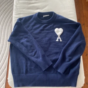 Mörkblå stickad tröja från Ami - Säljer en mörkblå stickad tröja från Ami Alexandre Mattiussi med deras ikoniska vita hjärta och A-logga på bröstet. Tröjan har rund halsringning, långa ärmar och breda ribbade muddar vid ärmslut och nederkant. Perfekt för en stilren och trendig look.