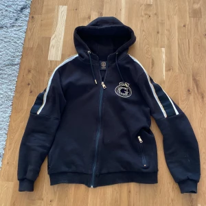 Svart hoodiejacka från Glorious Gangsta - Svart hoodiejacka med dragkedja från Glorious Gangsta. Jackan har guldiga detaljer, broderad logga på bröstet och vita ränder längs ärmarna. Justerbar huva med snörning och två fickor med dragkedja.